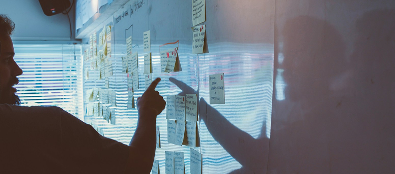 Pessoa a apontar para notas adesivas num quadro branco durante uma reunião de planeamento, com luz natural a entrar pela janela.